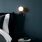 A black Clérachic wall light with a lit white glass globe sits above a bed with a dark upholstered headboard on a deep teal wall.