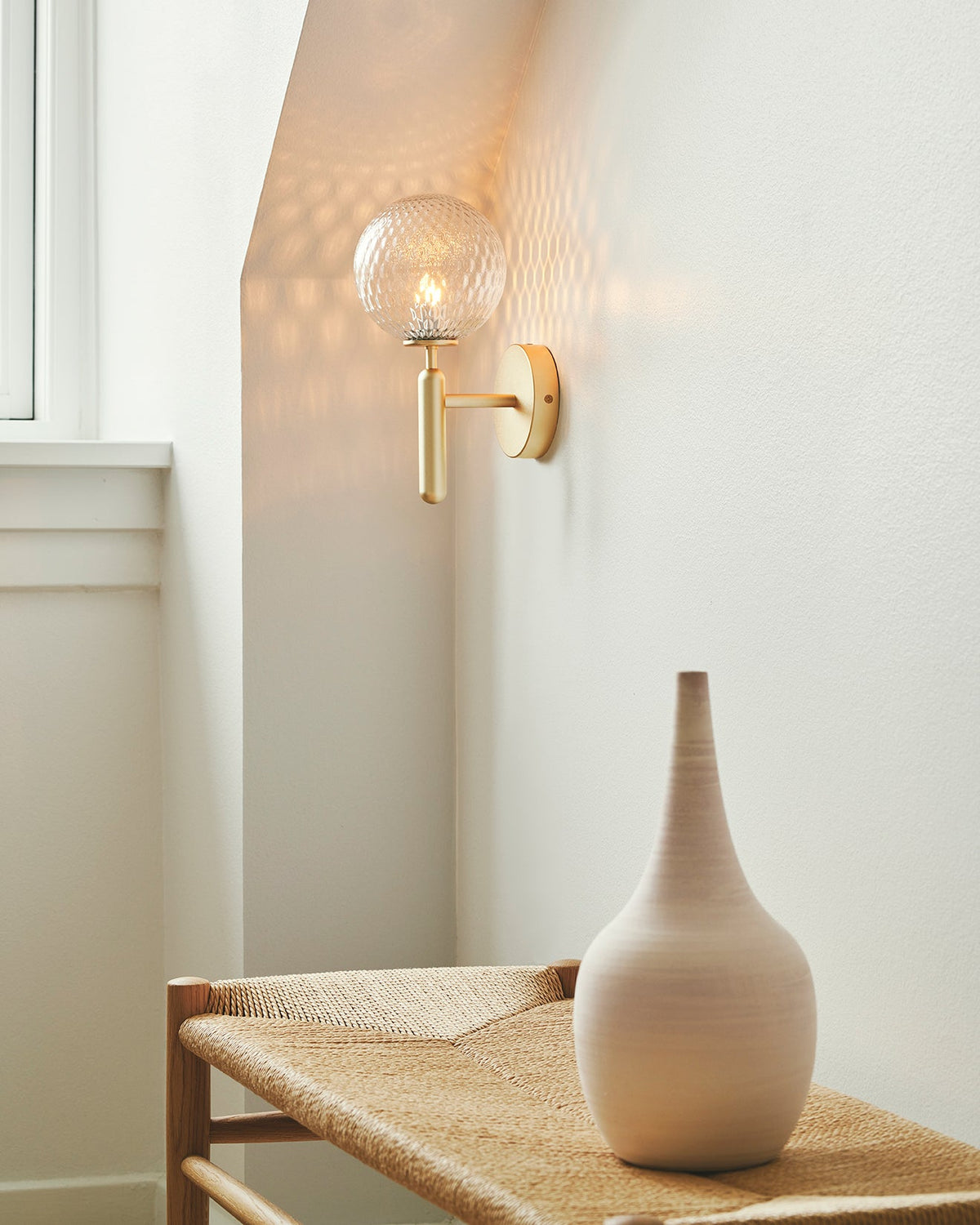 Near a window in an entryway, this brass Clérachic wall light's dimpled clear glass globe glows beside a woven bench and ceramic vase.