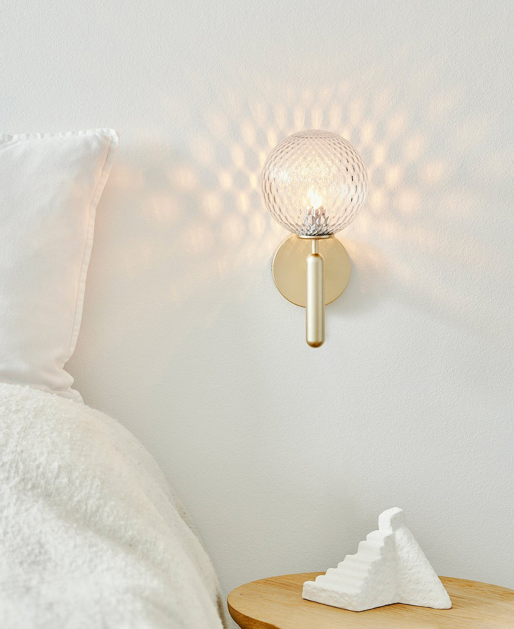 With a dimpled clear glass globe casting a honeycomb pattern of light, this brass Clérachic wall light sits above a bedside table.