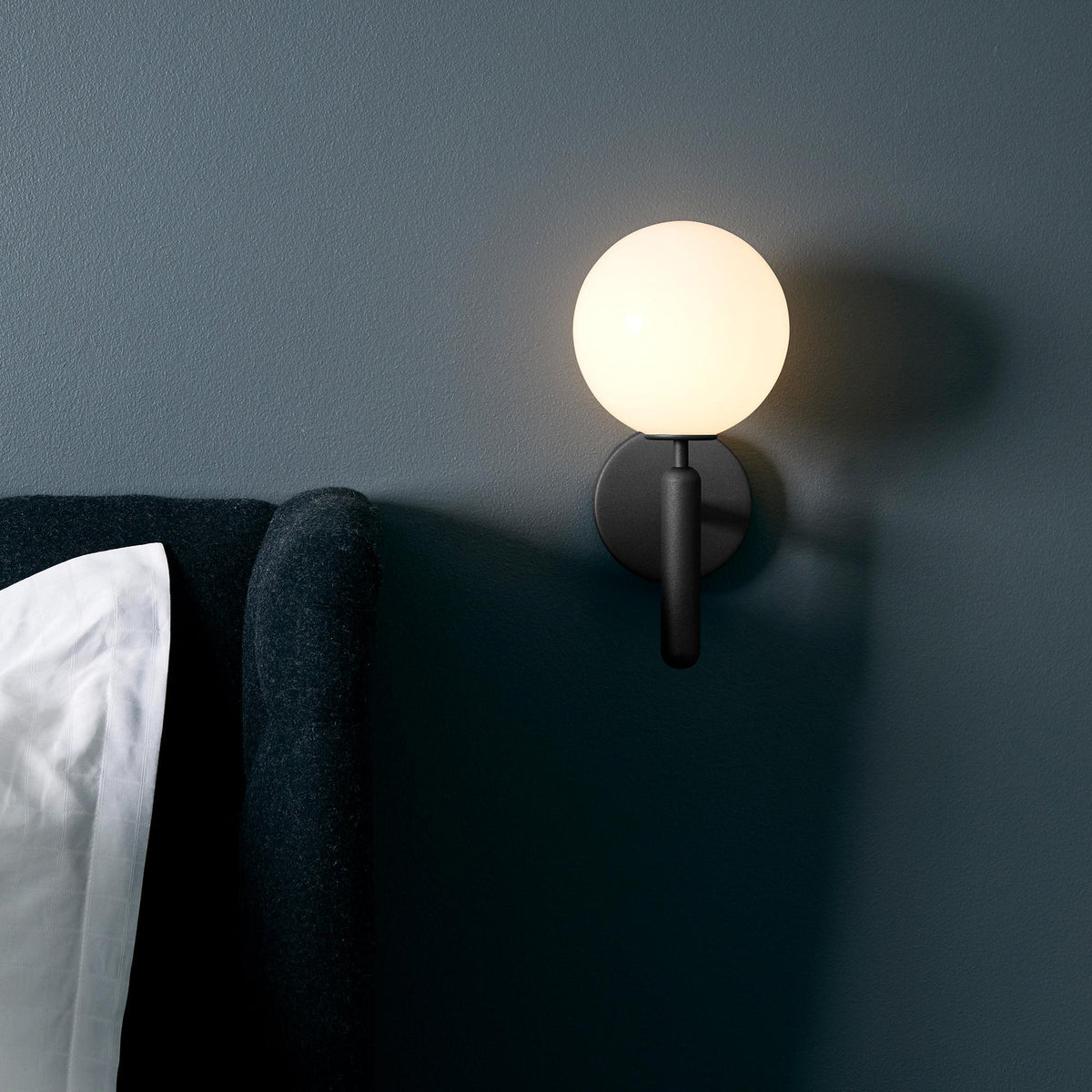 Above a bed with a dark upholstered headboard, this black Clérachic wall light glows on a deep teal wall.