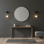 Black-finish Clérachic wall lights flank a round mirror above a wooden console table on a dark charcoal wall.