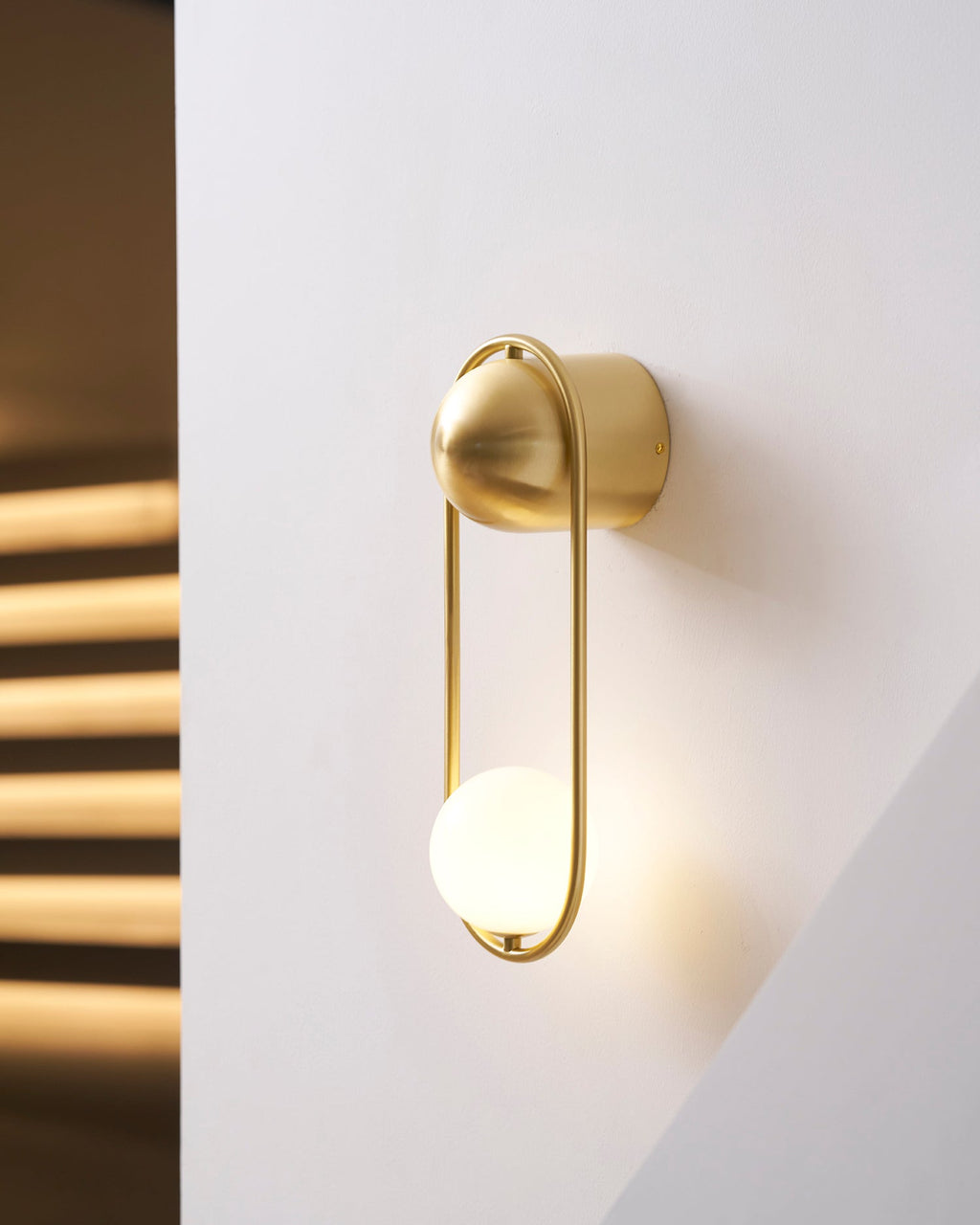 Close beside the sunlit stairwell, this brass Clérachic wall light glows warm against the streaked shadow on the wall.