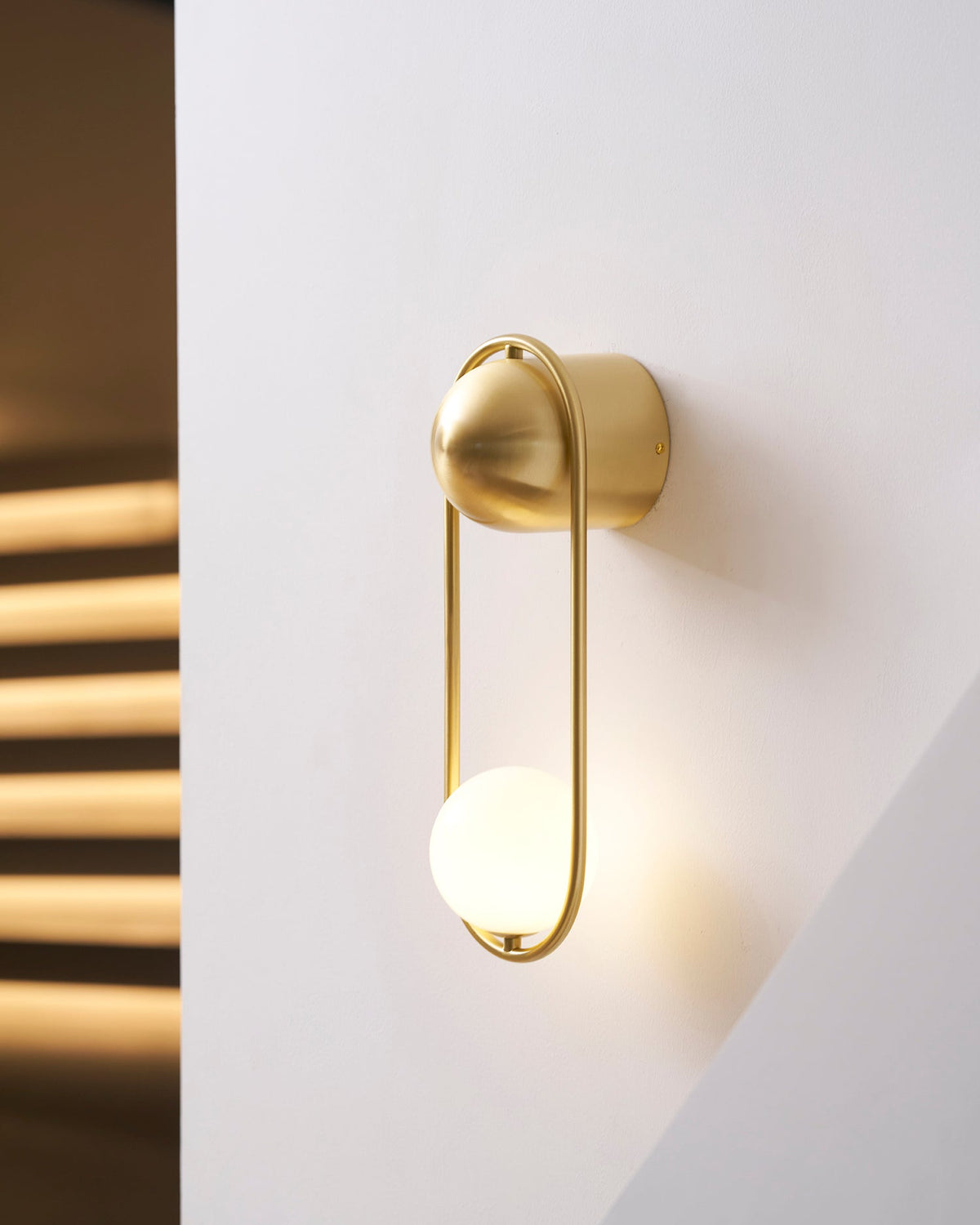 Close beside the sunlit stairwell, this brass Clérachic wall light glows warm against the streaked shadow on the wall.