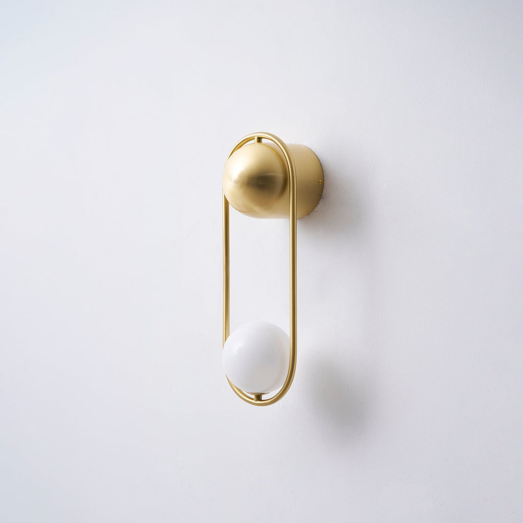 Angled view of this brass Clérachic wall light on a plain wall, its opal glass globe unlit and casting a soft shadow.