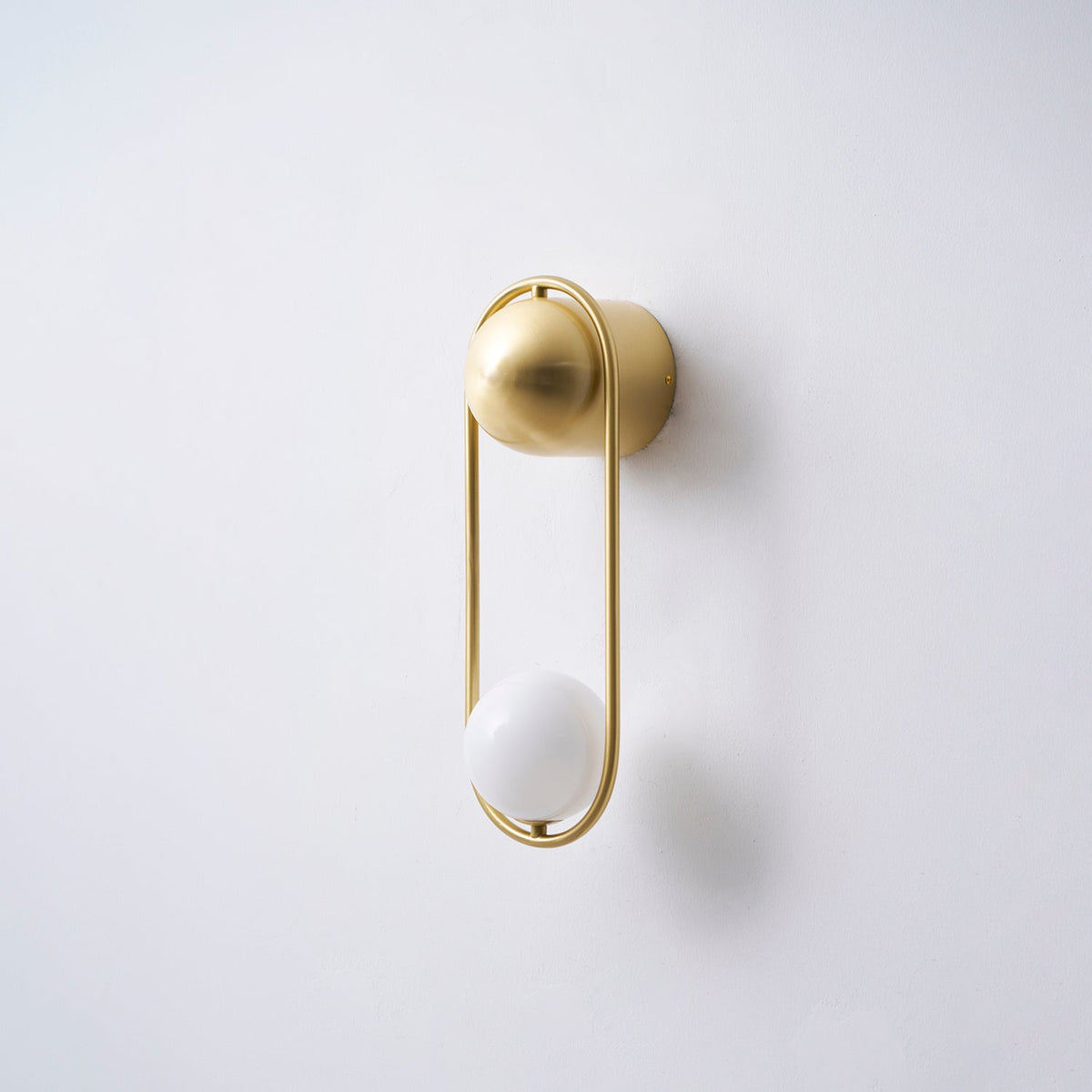 Angled view of this brass Clérachic wall light on a plain wall, its opal glass globe unlit and casting a soft shadow.