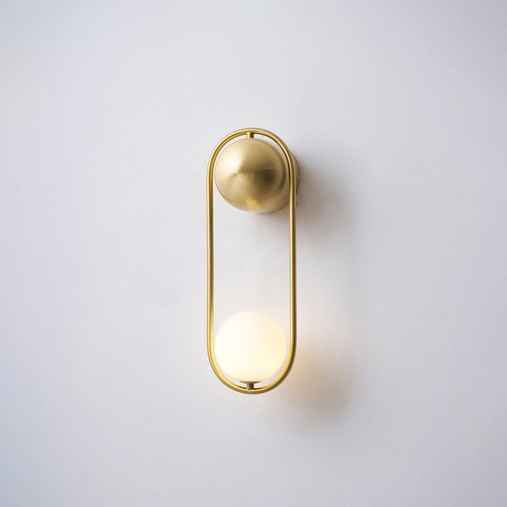 This brass Clérachic wall light on a plain wall, its opal glass globe glowing warm at the base of the loop.