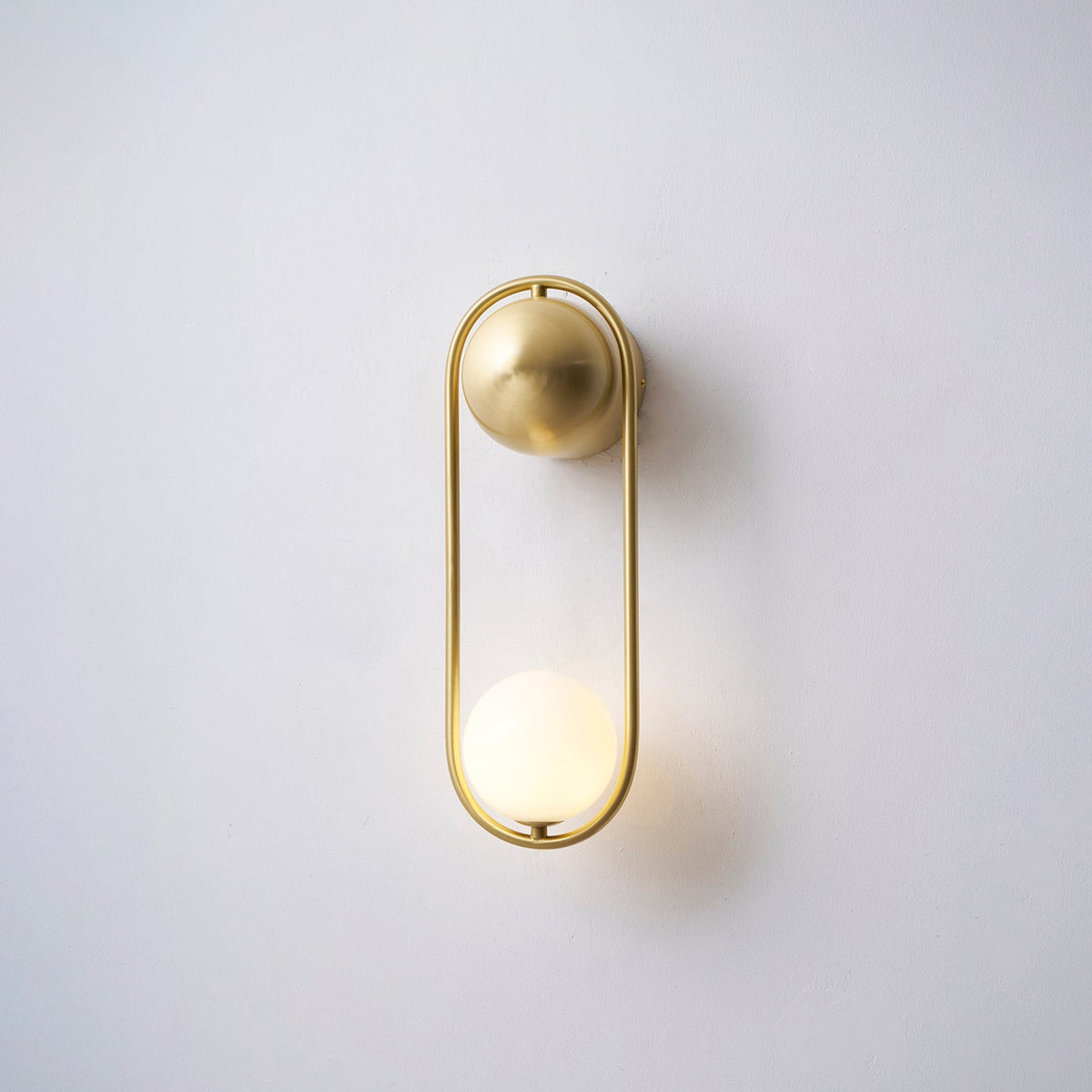 This brass Clérachic wall light on a plain wall, its opal glass globe glowing warm at the base of the loop.