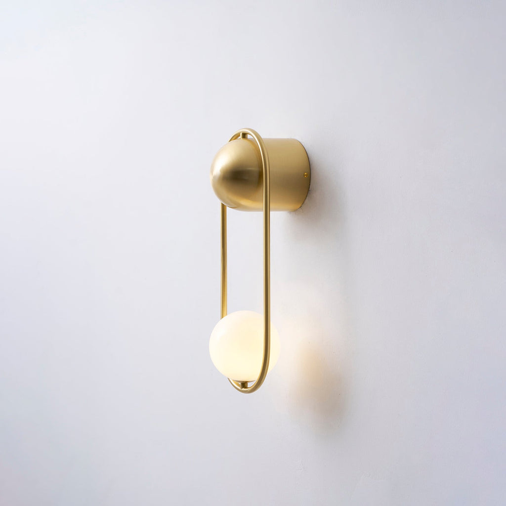 Angled view of this brass Clérachic wall light on a plain wall, its opal glass globe glowing warm.
