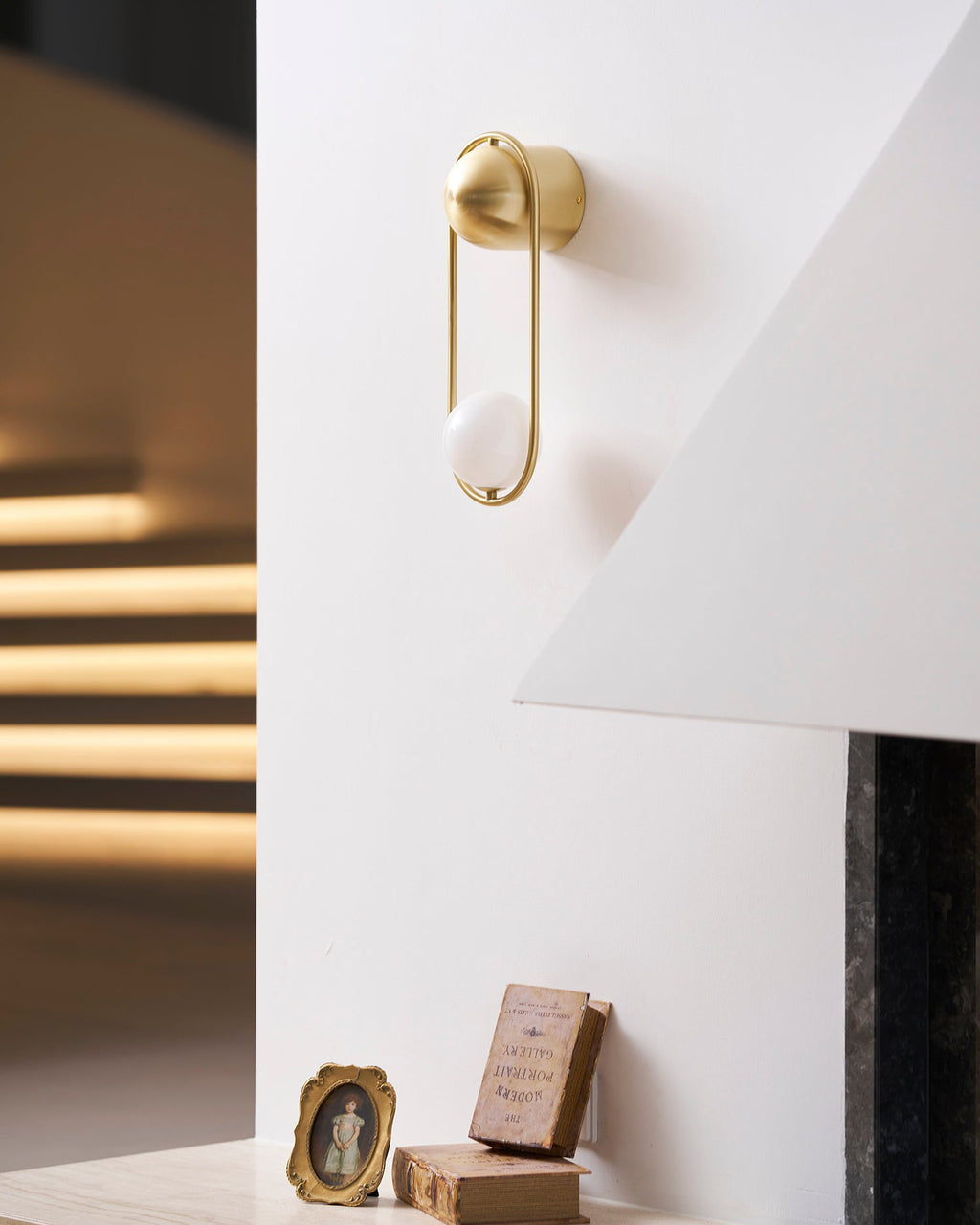 Near a staircase with sunlight streaking across the wall, this brass Clérachic wall light sits above a small shelf of books.