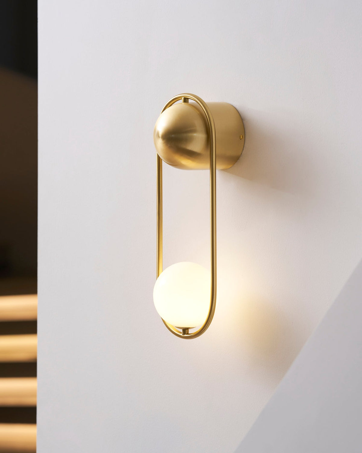 Against a wall with a dark triangular shadow, this brass Clérachic wall light glows warm through its opal glass globe.
