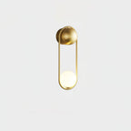 A brass Clérachic wall light on a plain white background, its opal glass globe glowing at the base of the loop.