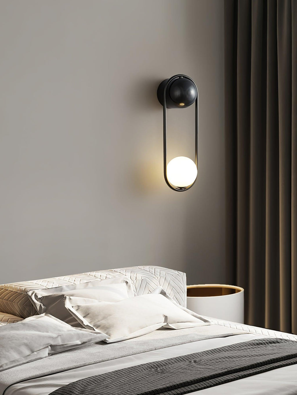 Above a patterned headboard beside dark curtains, this black-finish Clérachic wall light glows softly on a taupe wall.