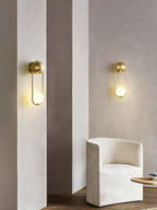 Two brass Clérachic wall lights sit on plaster pillars beside a cream armchair, shown unlit in daylight.