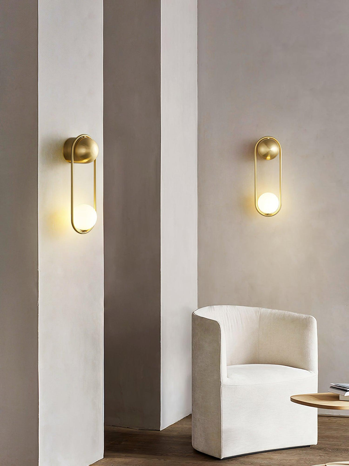 Two brass Clérachic wall lights sit on plaster pillars beside a cream armchair, shown unlit in daylight.