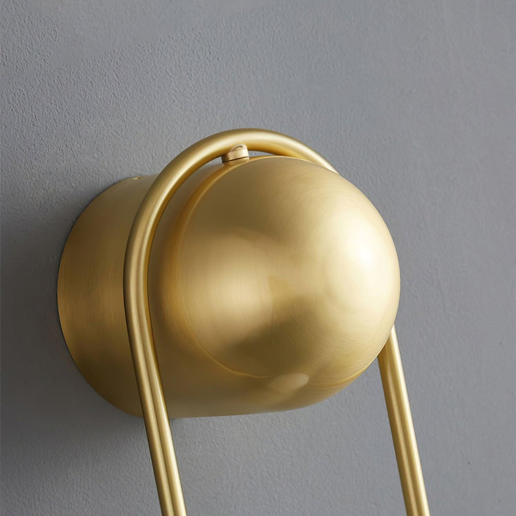 Extreme close-up of the brass ball top and curved loop on this Clérachic wall light against a plain grey wall.