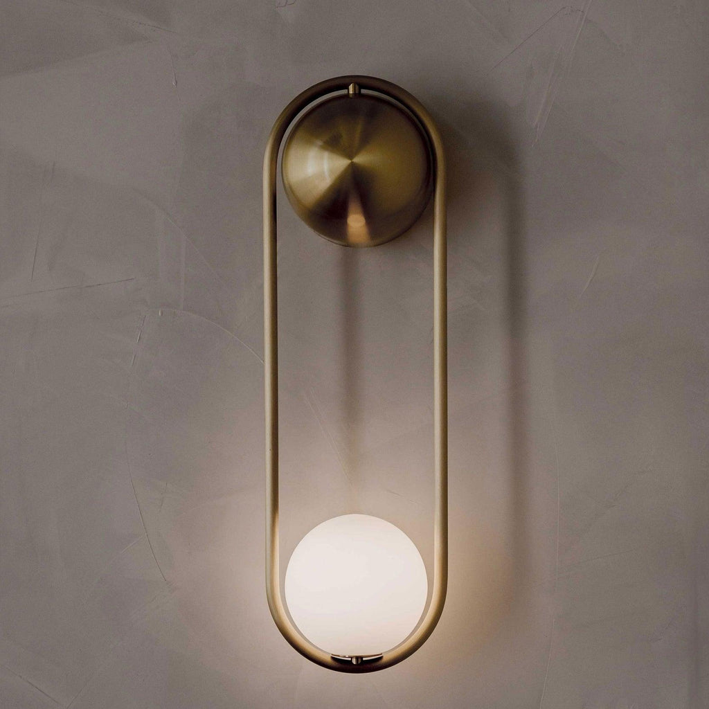 Close view of this brass Clérachic wall light on a textured plaster wall, its opal glass globe glowing warm at the base.