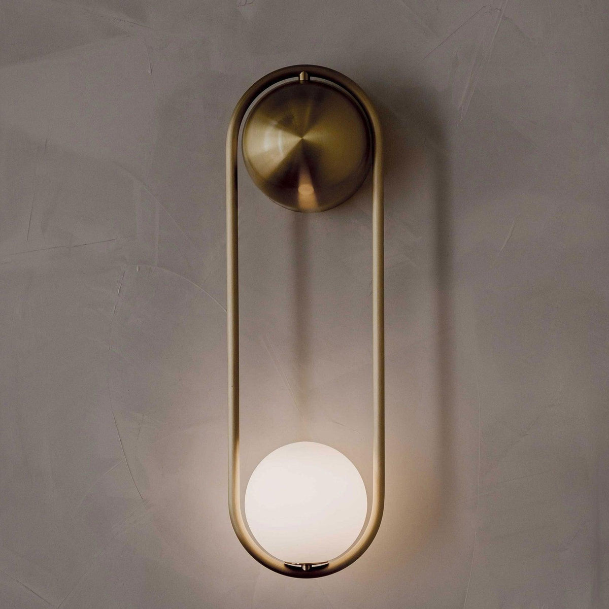 Close view of this brass Clérachic wall light on a textured plaster wall, its opal glass globe glowing warm at the base.