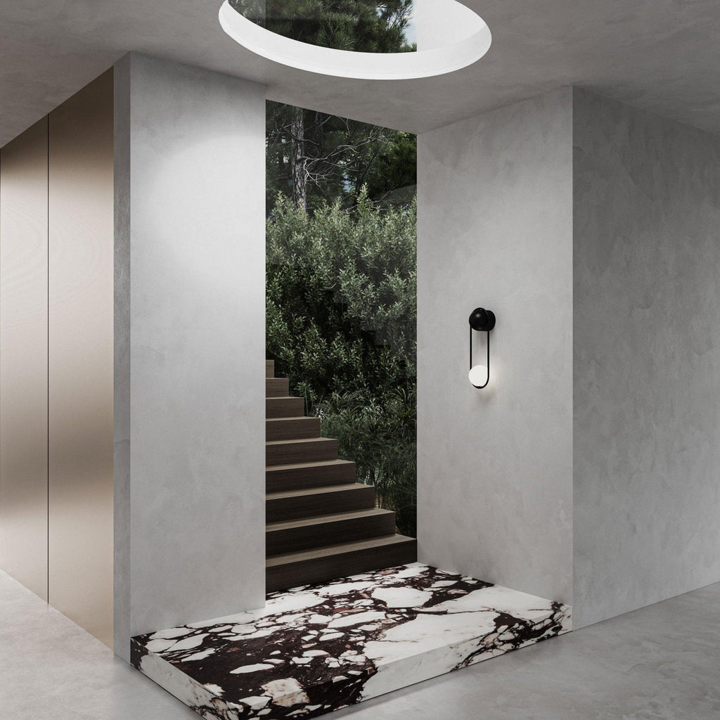 Beside an interior stairwell with greenery and a black-and-white marble step, this black-finish Clérachic wall light sits under a round skylight.