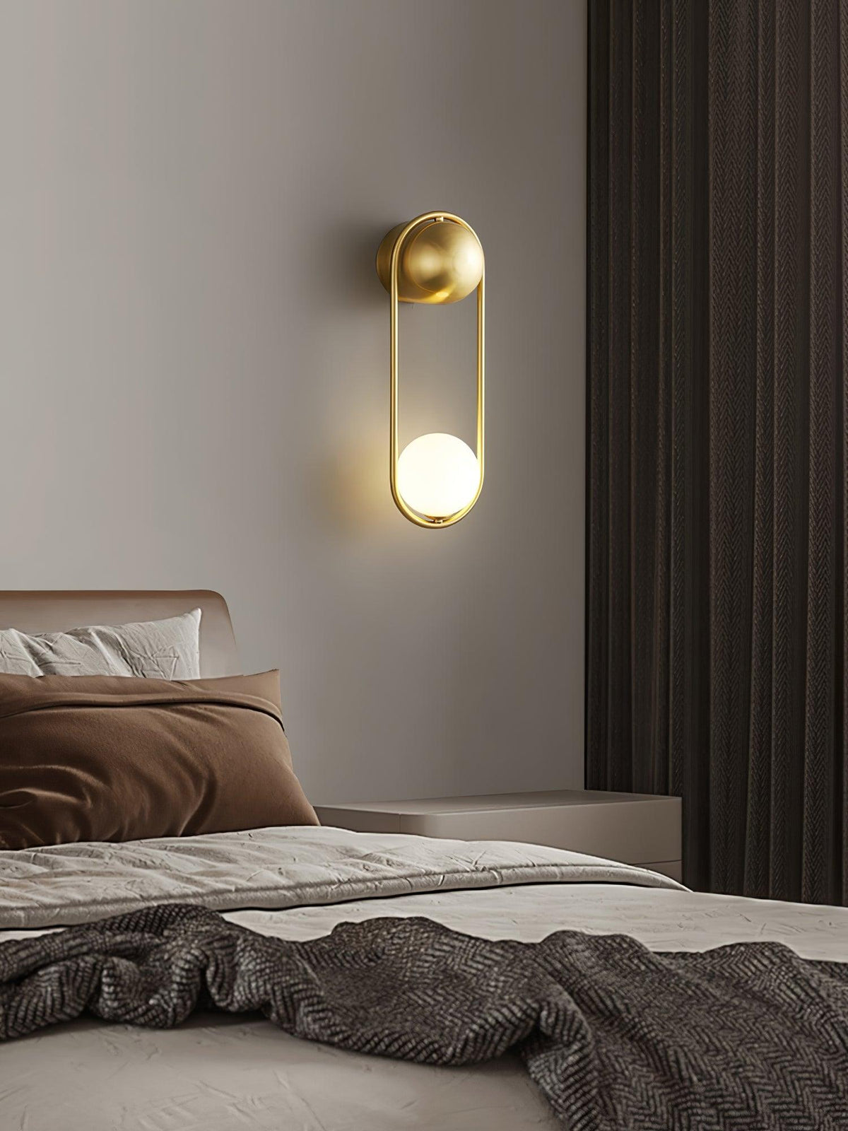 Above a bed with brown linens and dark curtains, this brass Clérachic wall light glows softly on a plain wall.