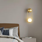 Above a camel-brown headboard beside a white nightstand with books, this brass Clérachic wall light glows softly.