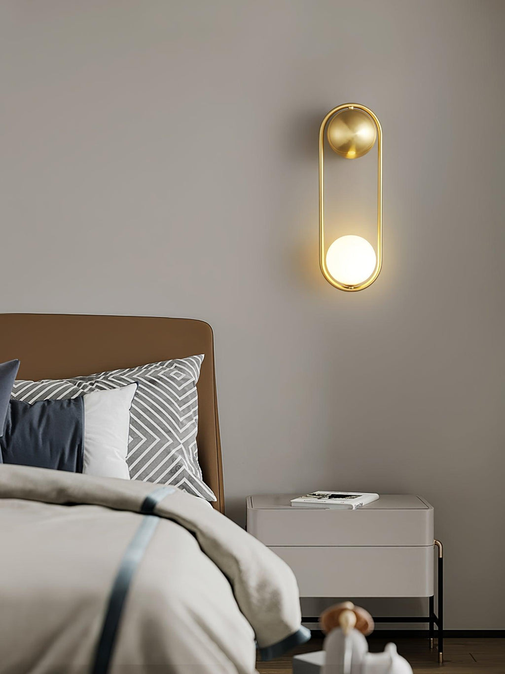 Above a camel-coloured headboard and a white nightstand, this brass Clérachic wall light glows softly on a taupe wall.