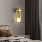 Mounted above a bed with brown linens and dark curtains, this brass Clérachic wall light glows through its opal glass globe.