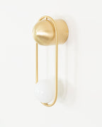 Side-angled view of this brass Clérachic wall light on an off-white background, its opal glass globe unlit.
