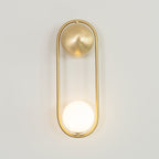Front view of this brass Clérachic wall light on a plain wall, showing its oval open frame and lit glass globe.