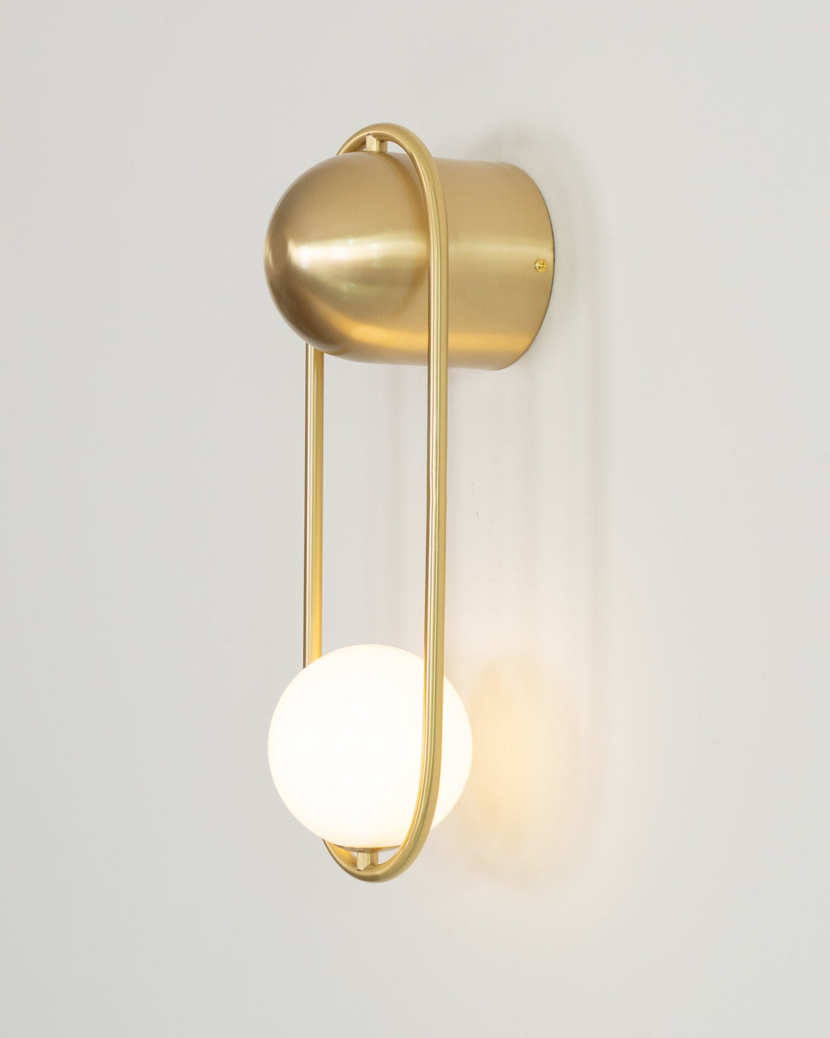 Angled side view of this brass Clérachic wall light on a plain wall, its opal glass globe glowing warm.