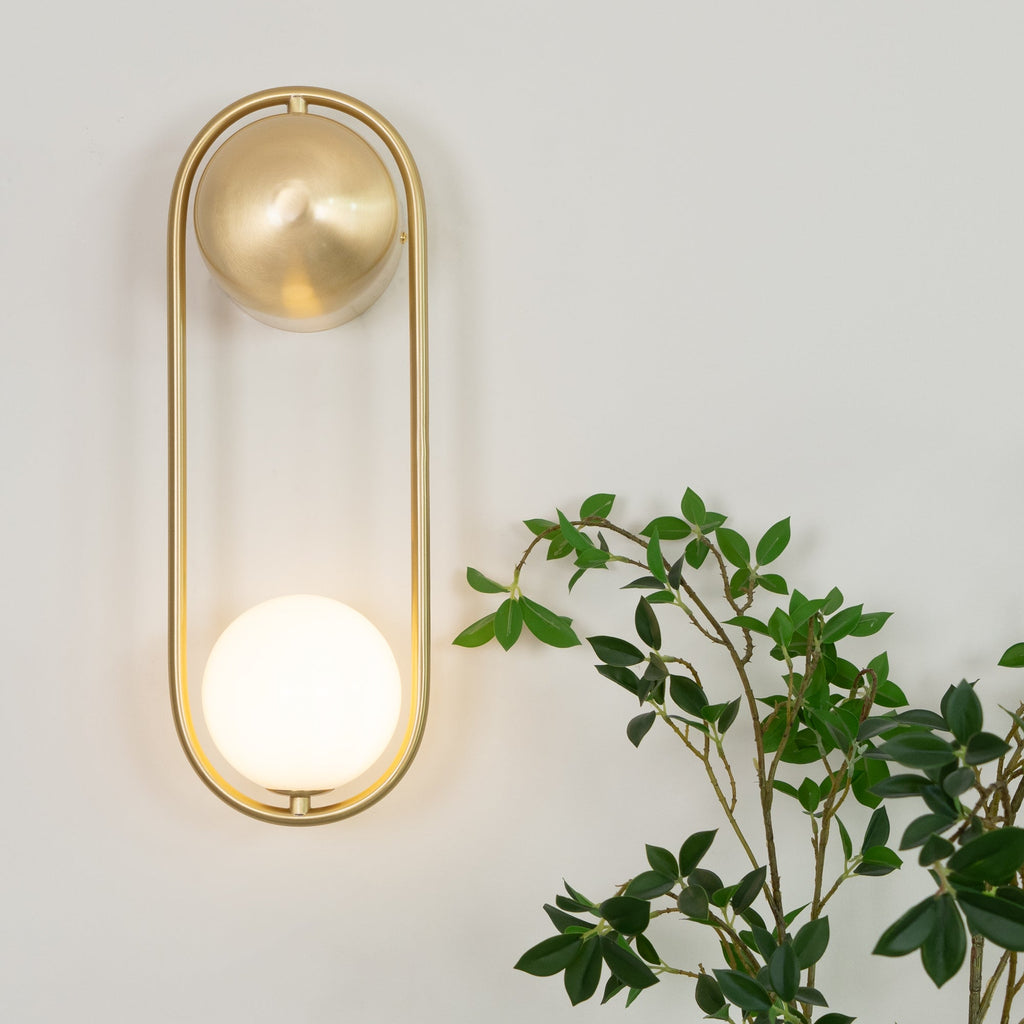 Beside trailing green leaves, this brass Clérachic wall light glows softly on a plain wall.
