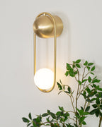 Close beside a leafy green plant, this brass Clérachic wall light glows warm against a plain wall.