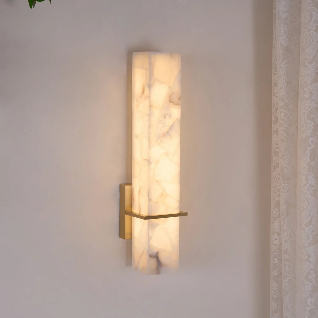 Alabaster cuboid wall light lit at an angle on a pale wall, brass crossbar catching the glow