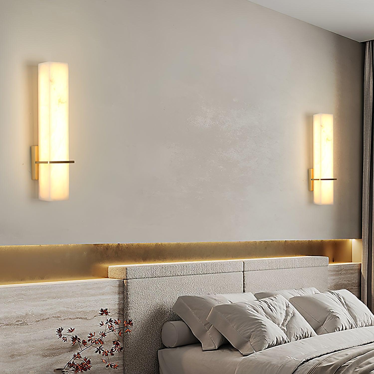 A pair of alabaster wall lights lit on either side of an upholstered bed headboard