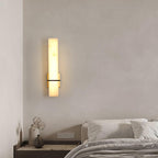 Alabaster wall light glowing beside a bed in white linen, above a nightstand of dried flowers