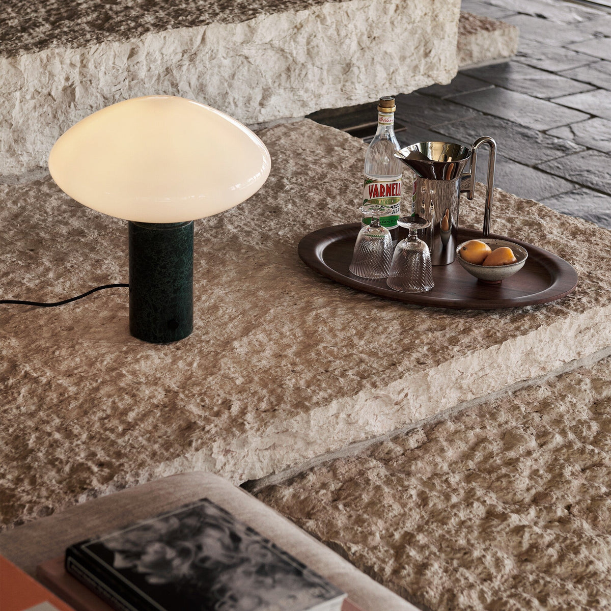 Clérachic table lamp lit on a rugged stone step outdoors, a bottle, glasses and a small bowl set on a tray beside it.