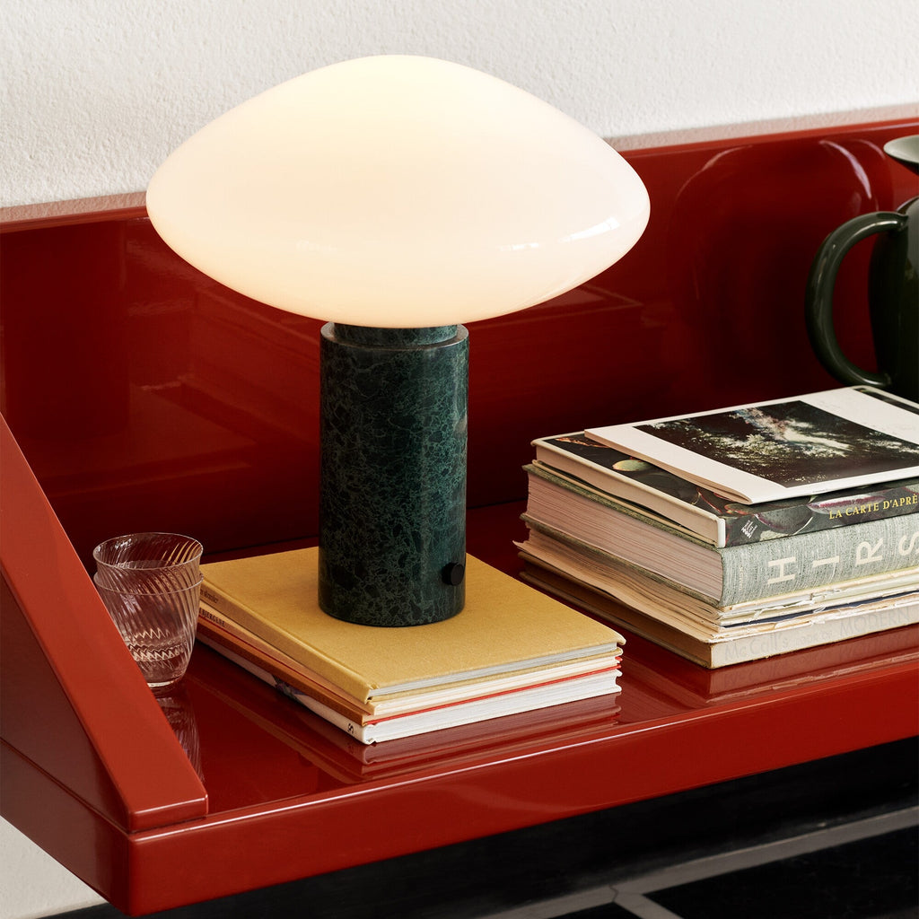 Clérachic marble table lamp lit on a red lacquered shelf, its glow set against stacked magazines and books.