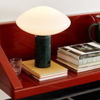 Clérachic marble table lamp lit on a red lacquered shelf, its glow set against stacked magazines and books.