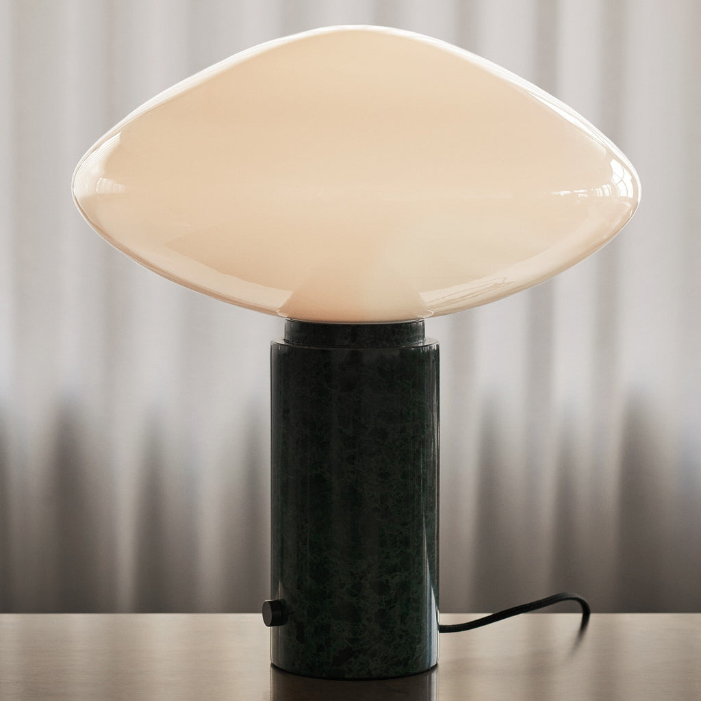Clérachic table lamp lit on a wood surface against curtains, a close view of the shade seam and green marble grain.