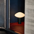 Clérachic table lamp lit within a concrete alcove, its glow set against a red floor and a deep-blue wall.