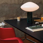 Clérachic table lamp lit on a black desk beside a red chair, a ceramic cup and books nearby, a framed print on concrete wall.
