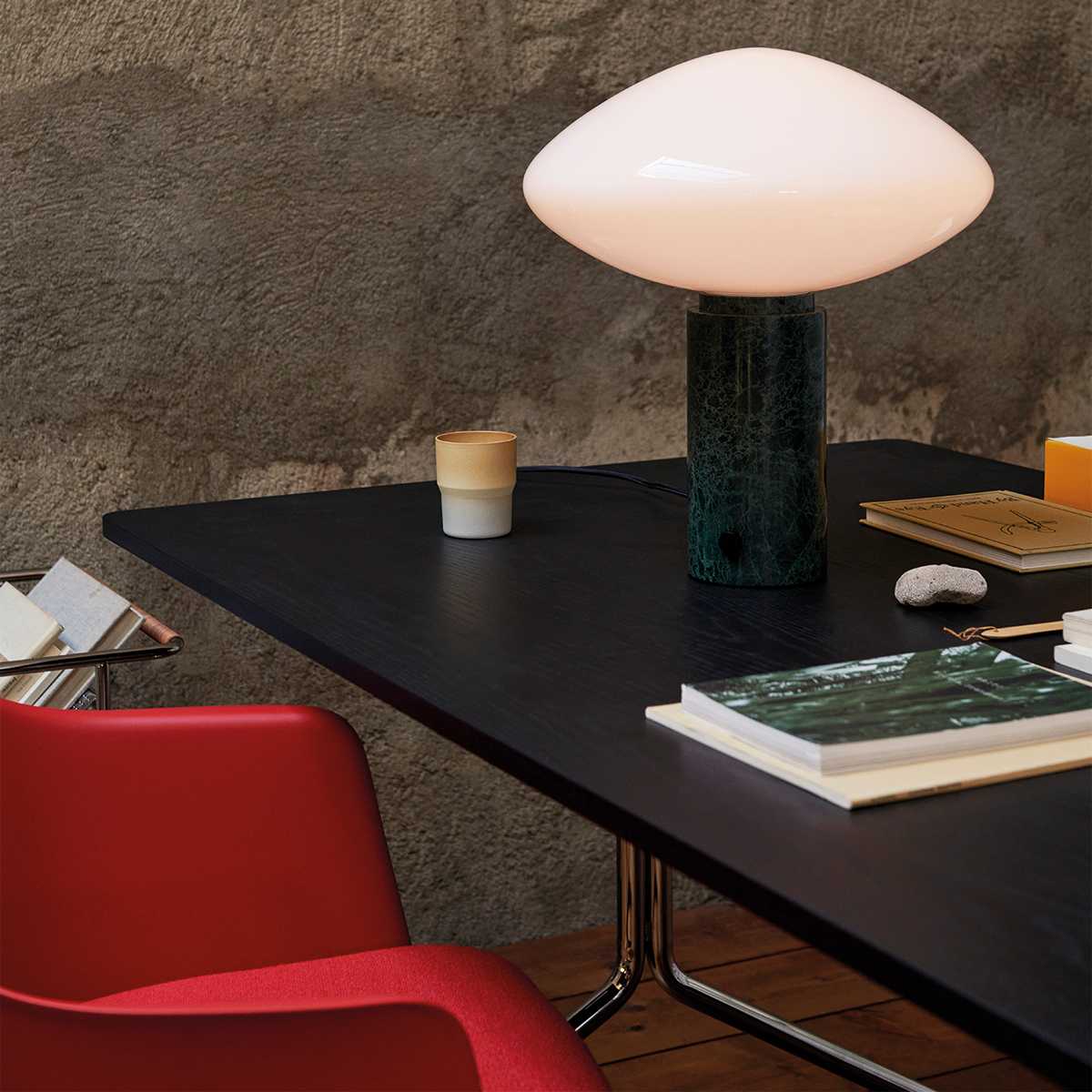 Clérachic table lamp lit on a black desk beside a red chair, a ceramic cup and books nearby, a framed print on concrete wall.