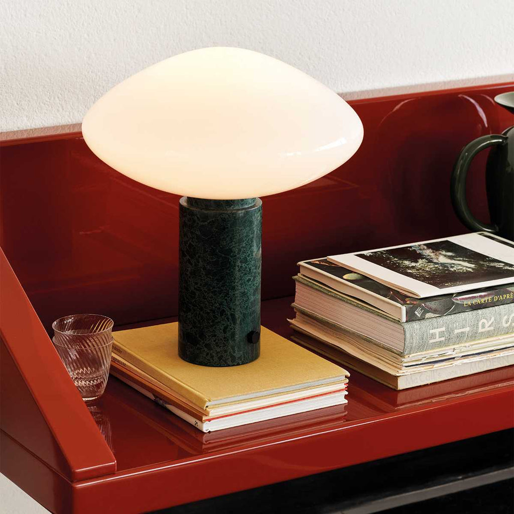 Clérachic marble table lamp on a glossy red shelf, a ribbed glass tumbler and layered magazines set beside it.