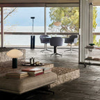 Clérachic table lamp on a low table beside stacked books, with glass doors open to a lake view behind it.