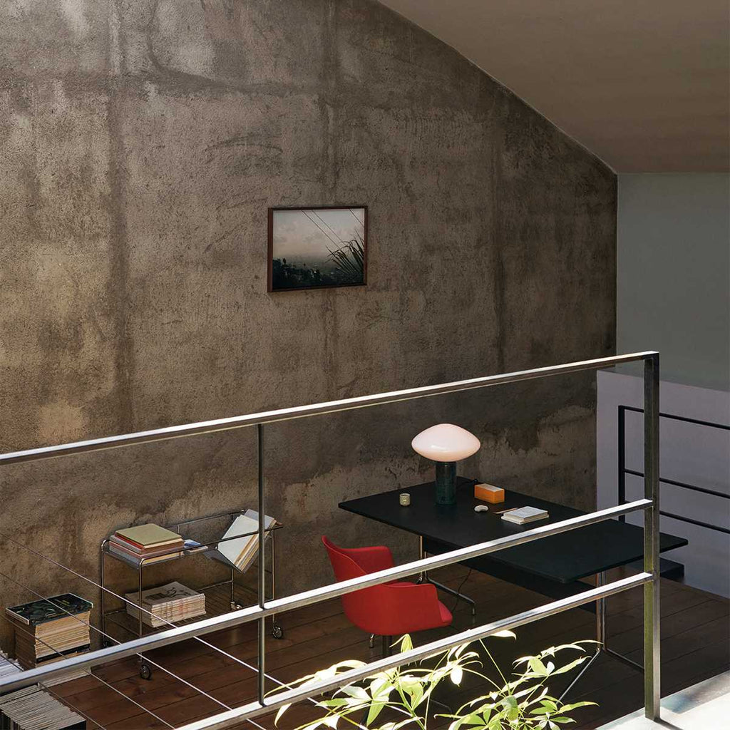 Clérachic marble table lamp lit on a black desk in a loft, seen past a glass balustrade and a red chair below.
