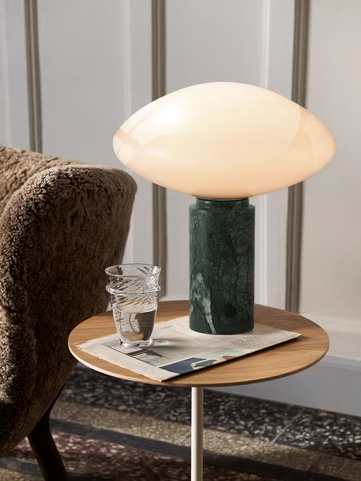 Clérachic table lamp lit on a round side table, cropped close on the stacked glass tumbler and the armchair's arm.