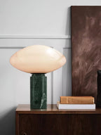 Clérachic marble table lamp lit on a dark wood sideboard, stacked books and a textured wall panel behind it.