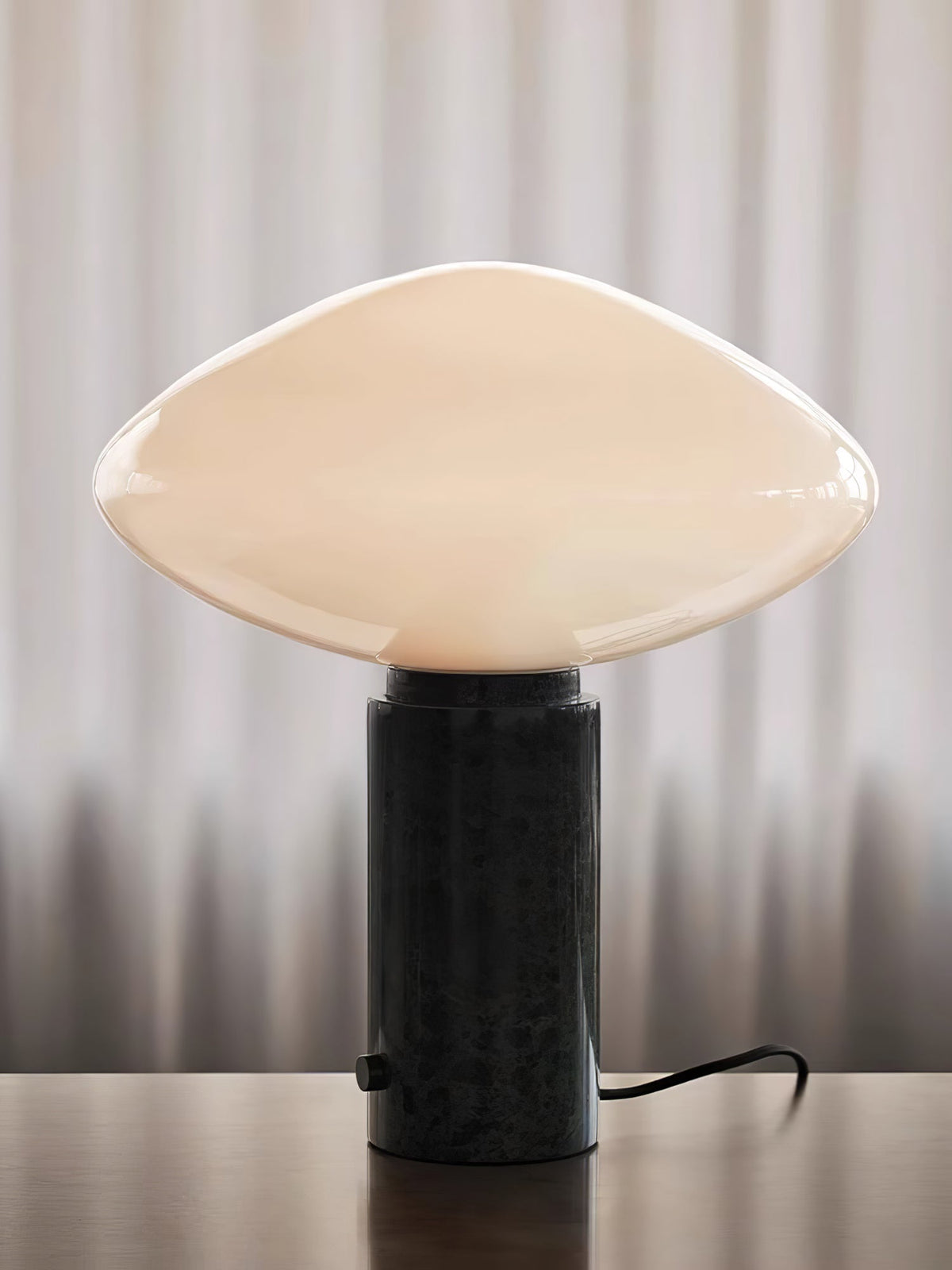 Clérachic table lamp lit against a curtained backdrop, its marble base thrown into deep shadow, cord trailing below.