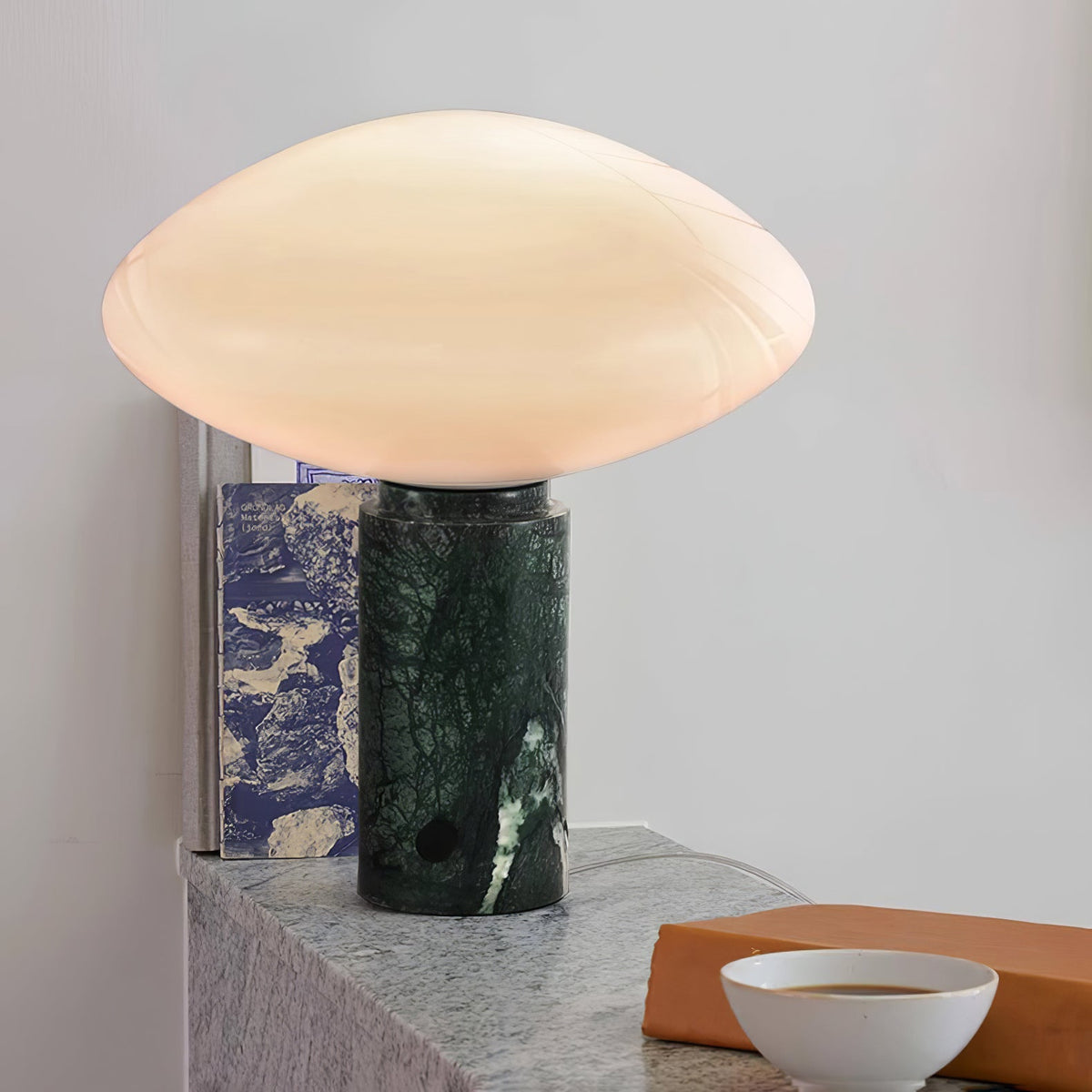 Clérachic table lamp on a pale stone ledge with a patterned blue book, a tan block and a white bowl nearby.