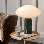 Clérachic table lamp lit on a round wood side table beside a bouclé armchair, a water glass and folded magazine nearby.