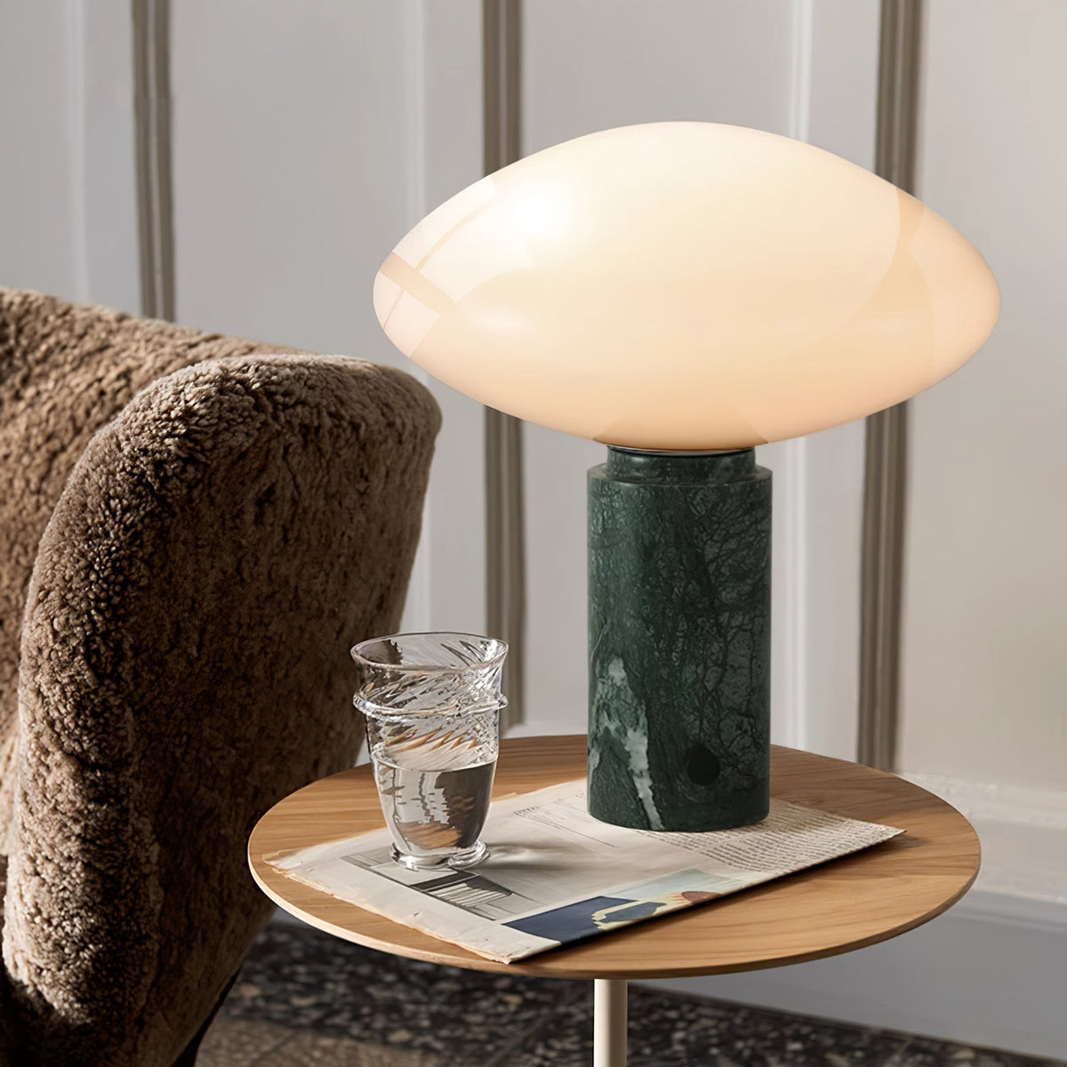 Clérachic table lamp lit on a round wood side table beside a bouclé armchair, a water glass and folded magazine nearby.